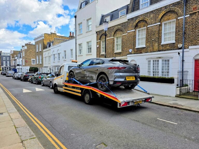 A UK flatbed recovery truck collecting a car from a fenced police impound compound at dusk