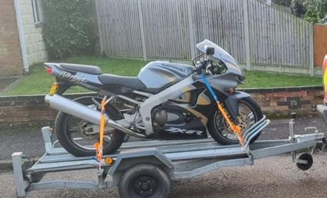 A motorcycle secured upright in a wheel chock on the deck of a UK flatbed recovery truck
